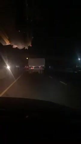The video shows a vehicle driving at night on a highway. In front of the vehicle, there is a large white truck, its taillights clearly visible. Above the road, an overpass casts a shadow over the scene. The road is divided by white lines, and the headlights of oncoming traffic are visible in the distance. The interior of the filming vehicle is dark, with only the dashboard slightly illuminated. The overall lighting is low, emphasizing the darkness of the night.