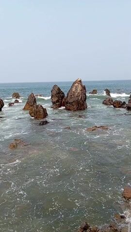 The video depicts a rocky shoreline where waves crash against dark, prominent rock formations. The rocks are scattered across the shallow water, creating a dynamic interplay between the solid land and the fluid sea. The water is a murky green, and the sky overhead is a pale, hazy blue. The scene is devoid of people and focuses primarily on the natural landscape.