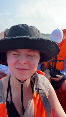 This video shows a group of four people on a red inflatable boat moving across a body of water. The person filming is in the foreground wearing a black bucket hat and an orange life vest. Behind them, a person in a blue shirt and an orange life vest looks down at their phone. Further back, a young person with long hair is visible, also wearing an orange life vest. On the far side of the boat, a person wearing a green hooded vest and an orange life vest is visible. The boat is moving, creating a wake. The environment appears to be a river or lake surrounded by trees under a bright sky. The video is shaky, suggesting it was filmed while the boat was in motion.