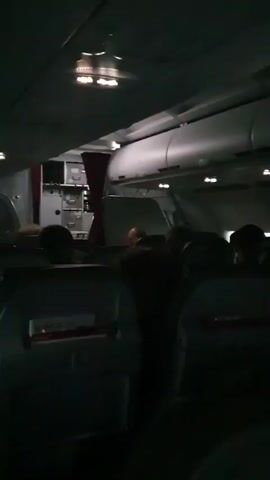 The video shows the interior of an airplane cabin. The lighting is dim, with some overhead lights visible. Rows of airplane seats are visible in the foreground and middle ground. Several passengers are seated in the chairs, their faces mostly obscured by the dim lighting and the angle of the camera. In the background, near the front of the cabin, a galley area is visible with cabinets and a red curtain. The overall impression is a dimly lit, typical airplane cabin scene during a flight.
