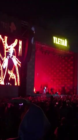 The video captures a vibrant outdoor concert scene at dusk in Lagunovo, Russia. The primary subjects are a large crowd of people gathered to watch a musical performance and the performers on a brightly lit stage.

The stage is a focal point, dominated by large screens displaying dynamic visuals. On the left screen, a close-up of a microphone with a fiery orange outline is visible. The main stage features a performer in a white shirt and dark pants, actively engaged in the performance. Behind the performer, the backdrop is a vibrant red, filled with a repetitive pattern that appears to be illuminated. Above the stage, a prominent yellow sign displays the word "YLETÄЙ" in bold capital letters. Stage lighting is a mix of pink and red hues, creating an energetic atmosphere. Several speakers and other stage equipment are visible.

The scene is bustling with activity. The crowd is dense, with many individuals holding up their phones, presumably to record or photograph the event. The overall mood appears to be energetic and celebratory, typical of a live music event. The time of day is evident from the dark sky, indicating either late evening or night. While weather is not explicitly shown, the outdoor setting suggests a clear night. The presence of a large crowd and stage setup clearly indicates a music festival or a large concert.