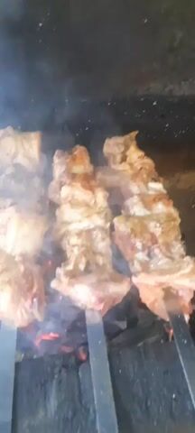 This is a close-up shot of meat skewers being grilled over hot coals. The meat appears to be chunks of chicken or pork, seasoned and browned. Smoke rises from the grill, obscuring some of the details. The skewers are arranged vertically, with several pieces of meat on each. The background is dark and indistinct, focusing attention on the cooking meat.