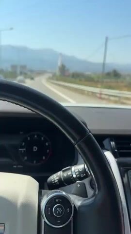 The video is a first-person view from inside a car driving on a highway. The steering wheel and dashboard are visible in the foreground, with a digital display showing the gear is in Drive and the current speed, 47 km/h. The road ahead is clear, with other cars in the distance. Mountains and hills are visible in the background under a clear blue sky. There are streetlights and roadside barriers along the highway. The navigation system on the car's infotainment screen shows a route to "Rio - Thira" with a distance of 4.7 km. There is also a notification for "Reduced speed" and "340 m".