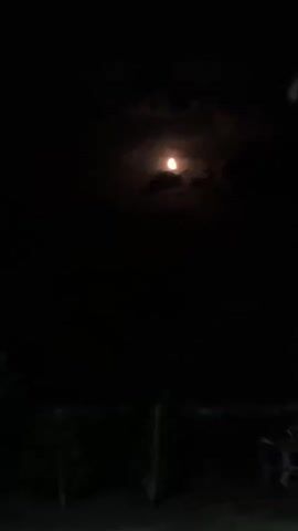 A video recording of a nighttime scene shows a bright moon partially obscured by clouds. The moon is positioned in the upper portion of the frame. The lower portion of the frame is dark, with silhouettes of vegetation and what appears to be fencing or structures visible. The scene is dark and appears to be outdoors.