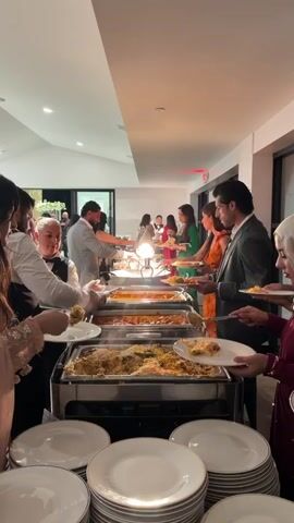 Several people in formal attire are lined up at a buffet, filling their plates with food from chafing dishes. A caterer in a black vest and white shirt is serving food from one of the dishes. There are multiple plates stacked at the front of the buffet, and the event appears to be indoors.
