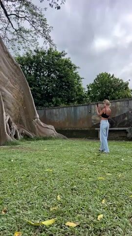 In a daytime outdoor shot, two women are present on a grassy lawn. To the left is an enormous tree with a pale, smooth trunk and complex root structure. A woman with dark hair, wearing a black tank top and dark pants, climbs onto the tree's roots, placing her hands on the trunk for support. 

To the right, another woman with blonde hair, dressed in a black top and light blue jeans, stands on the lawn and holds a phone up as if taking a picture of the woman climbing the tree. Behind her is a weathered, gray concrete wall with some patches of green vegetation. Trees with lush green foliage are visible above the wall.

The sky is overcast, creating a soft, diffused light across the scene. Fallen yellow leaves are scattered on the grass. The camera is positioned at a medium distance, capturing both women and the impressive size of the tree in relation to them.