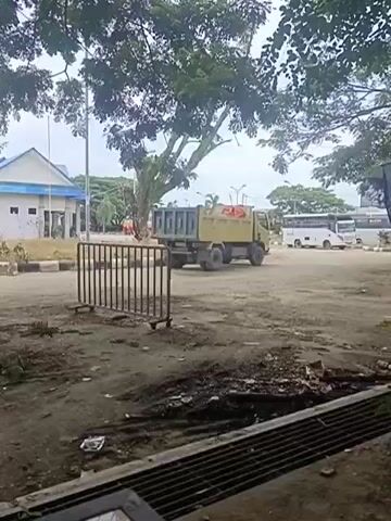 A dump truck is parked on a dirt lot, its bed is empty. In the background, there is a white building and several buses. Trees with green leaves are visible in the foreground and background.