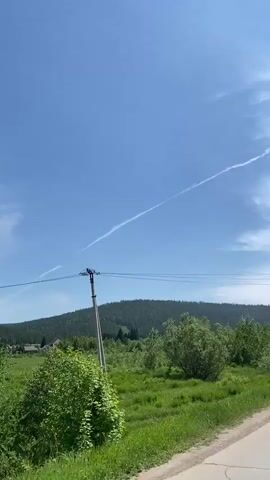 This is a first-person view of a car driving down a paved road. The sky is bright blue with a single white contrail stretching across it. There are power lines running horizontally across the frame, supported by a wooden pole on the left side. A dense, green forest covers a hillside in the background, with some trees closer to the road. The road itself is mostly out of focus.
