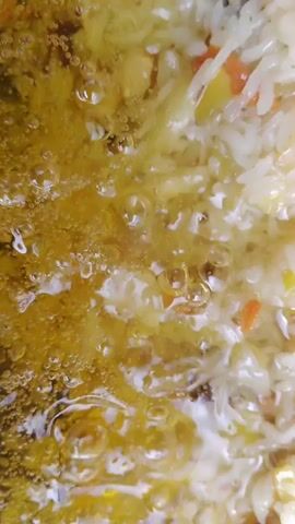 This is a close-up, overhead shot of pilaf being cooked in a large black wok. The rice is a mixture of white rice grains, shredded carrots, raisins, and chickpeas. The pilaf is partially submerged in a shimmering, golden oil on the left side of the wok. Several pieces of browned meat, likely lamb or beef, are interspersed throughout the rice. The wok is dark and appears to be made of cast iron, with some rust visible on the rim. The background is blurred, but hints of greenery suggest an outdoor setting. The camera slowly zooms out, revealing more of the wok and its contents.