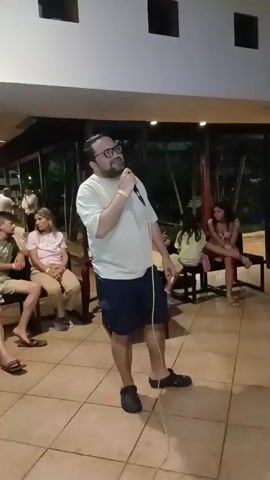 A man stands center frame holding a microphone and speaking into it. He is wearing a white t-shirt, blue shorts, and black Crocs. He has on glasses and a yellow wristband. He is standing on a tiled floor in what appears to be an indoor/outdoor covered area with a roof overhead. There are people seated on chairs in the background watching him. One woman in the foreground is wearing a white t-shirt and tan pants, and another woman behind her is wearing a light blue top and dark pants. To the right, a woman in a pink dress is seated on a bench. There is a light purple glow on the floor around the man's feet, suggesting stage lighting. A white cable runs from the microphone to the floor.