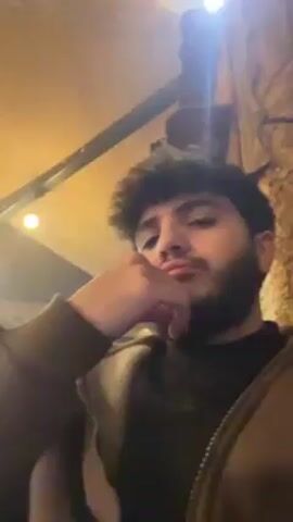 A young man with dark hair and a beard is in an indoor setting, possibly a café or restaurant. He is wearing a black hoodie and a brown jacket. He makes a peace sign with his fingers in the video. He holds a smartphone in one shot. The background has an artificial tent-like ceiling with several light bulbs. Another man is visible in the background, sitting in a chair. There appears to be some sort of smoke or vapor in the air. The camera is shaky and often points upwards.