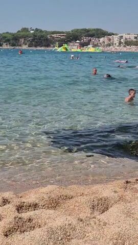 The video shows a sandy beach in the foreground with clear turquoise water. Several people are swimming and playing in the water. In the background, there is a coastline with buildings and vegetation. There is also a large inflatable water park in the sea. The sky is clear and blue, indicating a sunny day.