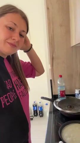 The video shows a person in a kitchen preparing food. A woman with long brown hair wearing a pink shirt stands to the left of the frame, touching her hair. In the background, a stovetop is visible with two frying pans. One pan contains a thin batter, likely for crepes or pancakes, and a lid rests on another pan. A plate of cooked crepes is on the right side of the stove. Various bottles and containers are on a shelf above the stovetop. The kitchen appears to have light-colored wooden cabinets.