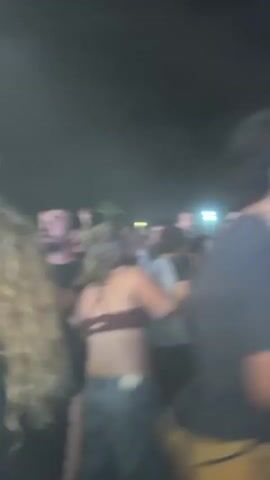 The video shows a crowd of people at night, likely at an outdoor event. Many people are dancing and singing along to music. The environment appears smoky.  People are dressed in casual clothing. A few raise their arms in the air, seemingly in excitement. The video is handheld and shaky.
