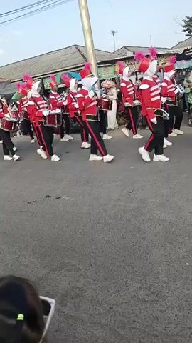 A marching band is parading down a paved road. The band members are wearing red jackets with black trim and accents, black pants, white shoes, and white head coverings with pink plumes on top. Many of them are carrying drums, some are holding instruments that resemble xylophones or glockenspiels. The band members are marching in formation. There are buildings and some trees visible in the background. Some spectators are standing on the side of the road watching the parade. The sky is overcast.