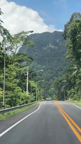 The video shows a paved road with double yellow lines and white dashed lines, winding through a lush green mountainous landscape. The road is bordered by a metal guardrail on the left and dense foliage on both sides. Numerous streetlights are visible along the left side of the road. In the distance, a large, verdant mountain dominates the skyline under a bright blue sky with scattered white clouds. A few vehicles are sparsely present on the road. The perspective suggests the video is taken from a moving vehicle.