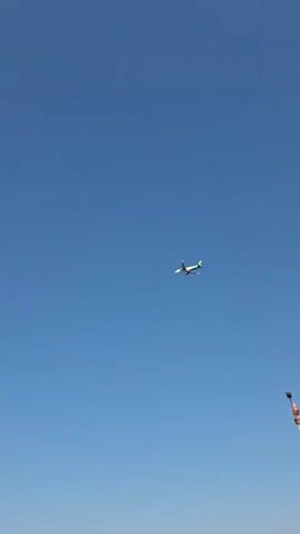 A small airplane with green accents flies across a clear blue sky. In the foreground, a highway with concrete barriers is visible. There are several streetlights along the highway, some of which have distinctive red and white patterned poles. A black SUV drives on the highway. The airplane is the primary focus of the video, while the highway and streetlights provide context and scale.