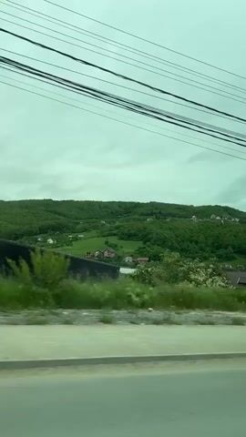 The video shows a passing view from a moving vehicle. The scenery consists of green hills with scattered houses, trees, and overgrown vegetation along the roadside. Overhead, several power lines are visible against an overcast sky. As the vehicle moves, it passes houses with tiled roofs.  The perspective suggests the camera is inside a vehicle, looking out the side window.