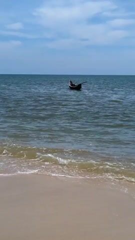 The video shows a tranquil scene at a beach. In the foreground, a sandy beach meets the gentle lapping waves of the ocean. The water is a deep blue, with a lighter, foamy crest on the waves closest to the shore. Further out in the ocean, a small, dark-colored boat is visible. A single person appears to be in the boat, holding what looks like an oar or a fishing pole. The sky above is a clear blue, dotted with a few white clouds. The overall impression is one of peace and solitude.