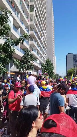 The video shows a large crowd of people gathered in what appears to be a protest or demonstration. Many are waving or wearing Venezuelan flags. The crowd is dense, with individuals of various ages and ethnicities visible. Some people are filming with their phones. The scene is outdoors, on a city street lined with tall buildings and trees. The sky is clear and blue. There is a light pole with many wires attached to it. A sign for a burger restaurant is visible. The atmosphere appears energetic and potentially politically charged.