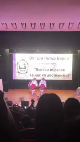 The video shows a performance on a stage with dancers in traditional Bulgarian clothing.  Four dancers are on stage, two in the front and two behind them, holding hands and performing a dance. Behind them is a banner reading "SU 'Dr. Petar Beron' gr. Cherven bryag", as well as the phrase "Vsichki vurkhove sa nas sa dostizhimi!'". The stage has a wooden floor. An audience is visible in the foreground, with heads and hands visible holding recording devices.  The lighting in the auditorium is dim, but the stage is well-lit. Above the stage is a row of decorative lighting fixtures. The video appears to have been recorded by a member of the audience.