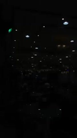The video depicts a dimly lit indoor space filled with people seated at tables. The lighting is provided by recessed ceiling lights and a chandelier, creating an ambiance of subdued illumination. Individuals appear to be engaged in conversation and dining. The darkness obscures most details, rendering the scene somewhat indistinct.