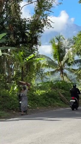 A person walks along a paved road carrying a bundle of wood on their head. A motorcycle is parked on the left side of the road. The surrounding area is lush with green vegetation, including palm trees and banana trees. In the background, a mountain can be seen against a blue sky with scattered clouds. The scene appears to be a rural setting in a tropical or subtropical climate.
