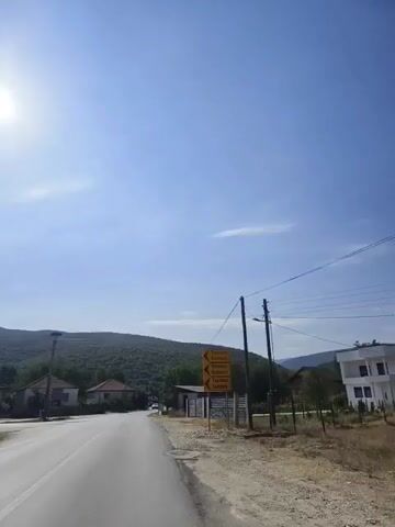 The video shows a view from a moving vehicle on a paved road. The road curves to the left, with a clear sky and sun visible. On the left side of the road, there are houses and a hill covered with trees in the background. A telephone pole with a bird's nest on top is also visible. On the right side of the road, there is a signpost with directional arrows pointing to different locations. The sign has text that appears to be in a foreign language. Further down the road on the right, there is a fence and some construction or storage structures. The sun is bright and casting shadows, suggesting it is daytime. There are power lines running overhead. A car is visible on the road ahead.
