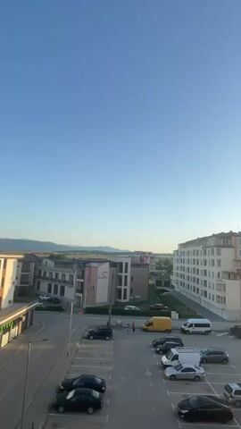 This is a vertical, medium-shot video of an urban scene, likely in Sevlievo, Bulgaria, during what appears to be late afternoon or early evening. The sky is a clear, bright blue, suggesting fair weather.

The scene is dominated by several multi-story buildings of modern design, some with balconies. In the foreground, a parking lot is filled with various vehicles, including sedans, SUVs, and vans. A yellow van is parked near the center of the frame, and a small, reddish-brown figure, possibly a person, is standing near it. Several cars are parked in the lot, with some appearing to be in motion or recently parked.

To the left, a lower commercial building with storefronts is visible, one with text that appears to read "APTEKA" (pharmacy in Bulgarian). Behind the commercial building, more residential buildings can be seen, with one displaying a sign with the word "Hotel" and "Palazzo" in its design. In the distance, rolling hills or mountains are silhouetted against the horizon, indicating a semi-rural or suburban setting.

The overall impression is one of a calm, ordinary day in a residential and commercial area. There are no overt signs of a specific event, but the presence of parked cars and the general ambiance suggest a typical scene in a town. The lighting indicates the sun is beginning to set, casting long shadows and bathing the scene in a warm glow.