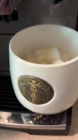 In the video, a Starbucks mug containing a few marshmallows and some sort of brown liquid, possibly coffee, is being filled with white liquid from an espresso machine. The mug is white and features a bronze Starbucks logo. Steam is rising from the mug as the white liquid is dispensed into it.