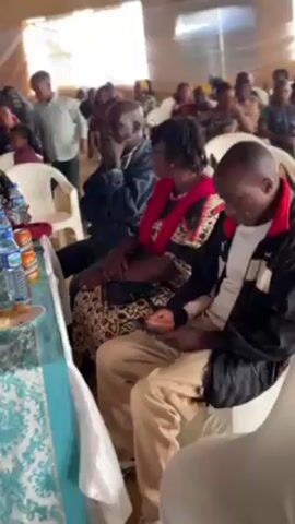 The video shows a medium shot of a room filled with people sitting at tables. The tables are covered in a blue and white patterned tablecloth and decorated with water bottles, food, flowers, and candelabras. People are wearing traditional African clothing and are seated in white plastic chairs. Some people are looking at their phones or interacting with each other.  The background is a beige wall with a white curtain.  The lighting is bright.