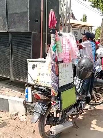 A motorcycle is parked next to a stack of large speakers. The motorcycle has a makeshift cargo carrier attached to the back, filled with various items including cups and what appears to be an umbrella. A man wearing a blue shirt and a cap is standing next to the motorcycle, interacting with someone off-camera. Another person in a blue shirt is visible behind him. The environment appears to be outdoors, with a building and other people in the background. The scene suggests a gathering or event is about to take place or has recently occurred, possibly involving music or entertainment, given the presence of the speakers.