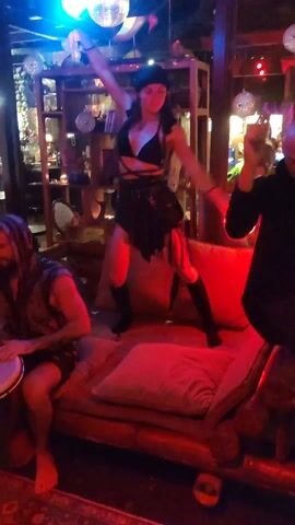 The video depicts a nighttime indoor setting, possibly a bohemian-style bar or lounge, where a woman is dancing atop a cushioned seating area. She's wearing a black outfit consisting of a bralette top, a wrap skirt with fringed detailing, and high boots. She manipulates two glowing orbs, one red and one blue, during her dance. To the left, a man is seated on the same furniture playing a hand drum or djembe. To the right, another man stands, seemingly recording the scene with a mobile device. The background reveals eclectic decor, including wooden shelves filled with various objects, a disco ball hanging from the ceiling, and ambient lighting. The overall atmosphere is festive and intimate.