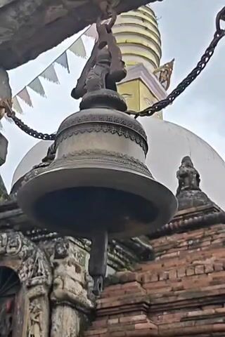 A large bronze bell is hanging from a chain, suspended from a wooden beam. The bell is ornate, with decorative patterns around its rim. It is positioned in front of a stupa-like structure, which is made of brick and stone. The stupa has a white dome and a conical spire at the top, with what appears to be a pair of eyes painted on it. Prayer flags, small and white, are strung between the beam and the stupa. The sky is overcast. The bell is stationary.