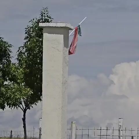 The video features a static outdoor shot centered on a tall, light-colored concrete pillar. A white stick or pole extends diagonally from the top right side of the pillar, with a piece of red fabric and a smaller piece of green fabric hanging from it. To the left of the pillar, a green leafy tree is visible. In the background, a metal wire fence stretches across the lower portion of the frame, and power lines are discernible at the top. The sky is partly cloudy with large white clouds, indicating a daytime setting. The fabric on the pole exhibits slight movement, suggesting a light breeze. No people are present, and no text is visible. A distinct, rhythmic percussive sound, resembling hammering, is audible throughout the clip. The scene is located in Ivan Vazovo, Bulgaria.