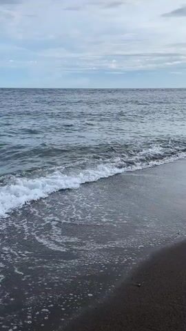 The video shows a close-up view of waves crashing on a dark sand beach. The water is a deep blue-gray, and the waves are small, creating white foam as they reach the shore. The sky is cloudy with patches of blue. The lighting suggests it is either early morning or late evening. There are no people or man-made objects visible.
