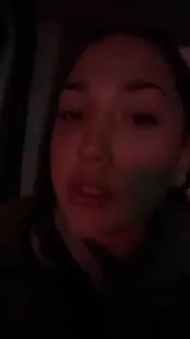 A young woman is singing into the camera while presumably sitting in a car. The car is dark, and the woman's face is illuminated by red light. She has dark hair and light skin. She appears to be wearing a sweater or jacket. The video focuses on her face as she sings and moves her mouth to the lyrics.