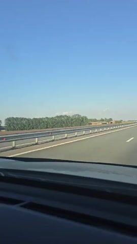 A first-person perspective video shows a highway with a clear blue sky and scattered clouds. A guardrail runs along the side of the road, separating the highway from a dense line of green trees. The road is paved and has white lane markings. The video captures the view from inside a vehicle, with the dashboard visible in the foreground. A light-colored car can be seen driving in the distance on the highway. The scene suggests a clear and sunny day, likely in a rural or suburban area.