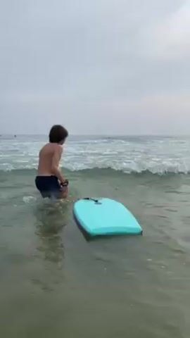 A young boy is standing in the ocean, facing away from the camera. He is wearing blue swim trunks and has short, curly brown hair. He is holding a leash connected to a light blue boogie board that is floating in the water. The water is shallow and clear, with small waves breaking in the distance. The sky is overcast and gray. The boy walks a few steps further into the water, pulling the boogie board behind him. He then bends down and touches the boogie board.