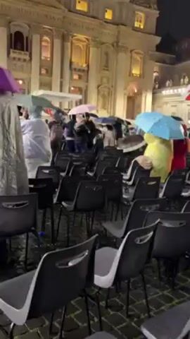 Here is a description of the video:

The video shows a nighttime outdoor scene of a crowd of people in Vatican City, apparently waiting for or attending an event in St. Peter’s Square.  Many people are holding umbrellas or wearing ponchos to shield themselves from the rain. Rows of empty black chairs are set up in front of the crowd. The architecture of St. Peter's Basilica is visible in the background, illuminated with lights. The statue of St. Peter is seen standing above the crowd in the distance.