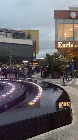 A public outdoor plaza features a fountain in the foreground with illuminated lights in the water. In the background, several businesses are visible. One building has a sign that reads "Earls Kitchen." People are walking, standing, and sitting at tables in the plaza. The sky is overcast, suggesting it may be evening or a cloudy day. The overall scene depicts a lively commercial area.