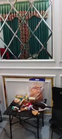 The video shows an indoor scene with a man sitting on a chair and looking at his phone. In front of him, there is a table with various items on it, including food, snacks, and a remote control. In the background, there are curtains, a mirrored wall with a diamond pattern, and a wall decoration that spells out "HOME" with a clock. The man appears to be relaxed, and there are no other people visible in the immediate vicinity. The lighting suggests it is daytime, and the overall atmosphere seems casual.