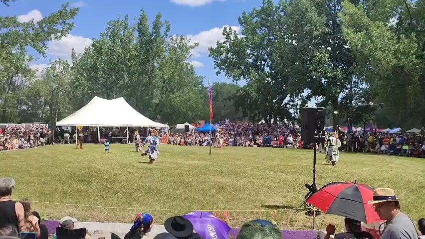 A wide-angle, slightly tilted shot captures an outdoor gathering on a sunny day, likely a powwow or festival. In the foreground, out-of-focus individuals are visible, some holding umbrellas. The middle ground features a large, cleared grassy area where several people are dancing. One individual in traditional regalia, adorned with feathers and colorful fabric, is performing a dance with energetic movements. Another person in similar attire stands to the right, facing the dancers. A large white tent is set up on the left, with people gathered underneath it. Behind the dancers, a crowd of spectators sits on bleachers and stands, watching the performance. Colorful flags are visible on poles, and trees line the background, suggesting a park or open field setting. A large speaker is positioned on a stand to the right of the dancing area. The overall atmosphere appears festive and engaging.
