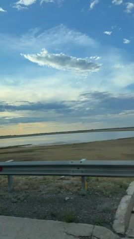 This video shows a scenic view of a body of water and the sky. The scene is set during the day, with a bright blue sky filled with fluffy white clouds and some darker, more dramatic clouds towards the horizon. The water appears calm and expansive, reflecting the sky. The foreground features a silver metal guardrail, typical of a roadside, and dry, sparse vegetation. In the distance, the landscape is flat and appears to be an open, possibly rural area. There are some distant structures visible on the horizon, and the overall impression is one of open space and natural beauty. The location is identified as Torghai Station, Kazakhstan. The video seems to capture a moment of travel, perhaps from a vehicle, as indicated by the perspective and the guardrail. The mood is peaceful and serene, evoking a sense of the vastness of the natural world. There is no visible text in the video.