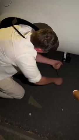 A person in a white shirt and light-colored pants is kneeling on the ground next to a white vehicle. The person is using a silver tool to operate a jack mechanism attached to the underside of the vehicle. The tool is being turned, implying the vehicle is being lifted or lowered. The ground appears to be asphalt. In the foreground, a hand is holding a smartphone, displaying a timer that reads 00:10 and other notification icons. The lighting suggests it is nighttime or a poorly lit area.