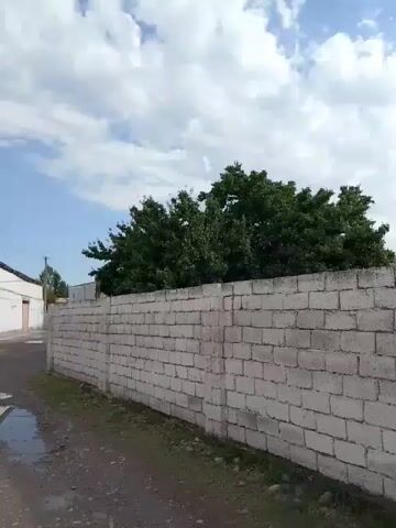 A video pans from left to right along a white brick wall. Behind the wall are green trees. Above the wall are white clouds against a blue sky. On the left side of the video, part of a white building and a dirt road are visible.