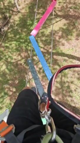 This is a first-person point-of-view video of someone navigating an aerial obstacle course. The subject is wearing black shorts, black Crocs with a floral pattern, and a harness with carabiners. The obstacle consists of a series of suspended, colorful planks – red, pink, and blue – that are staggered, requiring the person to step from one to the next. The ground below is covered in green grass and dappled with sunlight filtering through trees, suggesting a daytime setting in a park or forest. The overall mood appears adventurous and focused, as the subject carefully moves across the course. There is no visible text in the video. The location context provided is Lâm Viên - Đà Lạt, Vietnam.