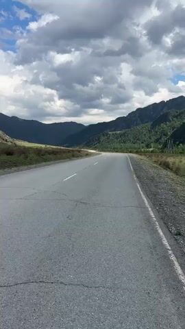 The video shows a scenic view of a mountain road. The road is gray asphalt, with white lane markings and some cracks. On either side of the road is low, green vegetation. In the distance, there are mountains. The sky is partially cloudy with patches of blue. The camera pans to the left, focusing on a rocky outcrop on the mountainside. The overall environment suggests a rural, mountainous area.