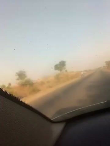 The video shows a view from inside a vehicle driving on a narrow asphalt road. The landscape is dry and hazy, with brown grass and sparse trees on either side of the road. A motorcyclist wearing a white shirt is seen in the distance, briefly. The sky is pale blue with a slightly dusty appearance. The camera angle is from the dashboard of the car, showing a portion of the dashboard. The video appears to be taken during the day. There is background music playing.