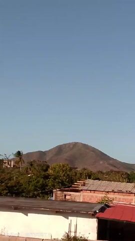 The video shows a view of a town or village with mountains in the background. The sky is mostly clear and blue with some white clouds appearing later in the video. The camera pans slowly across the scene, revealing buildings with tiled roofs, trees, power lines, and a street light. The mountains are covered in vegetation and appear somewhat dry.  The text "Infinix SMART8" is visible in the lower left corner of the screen, indicating the device used to record the video.